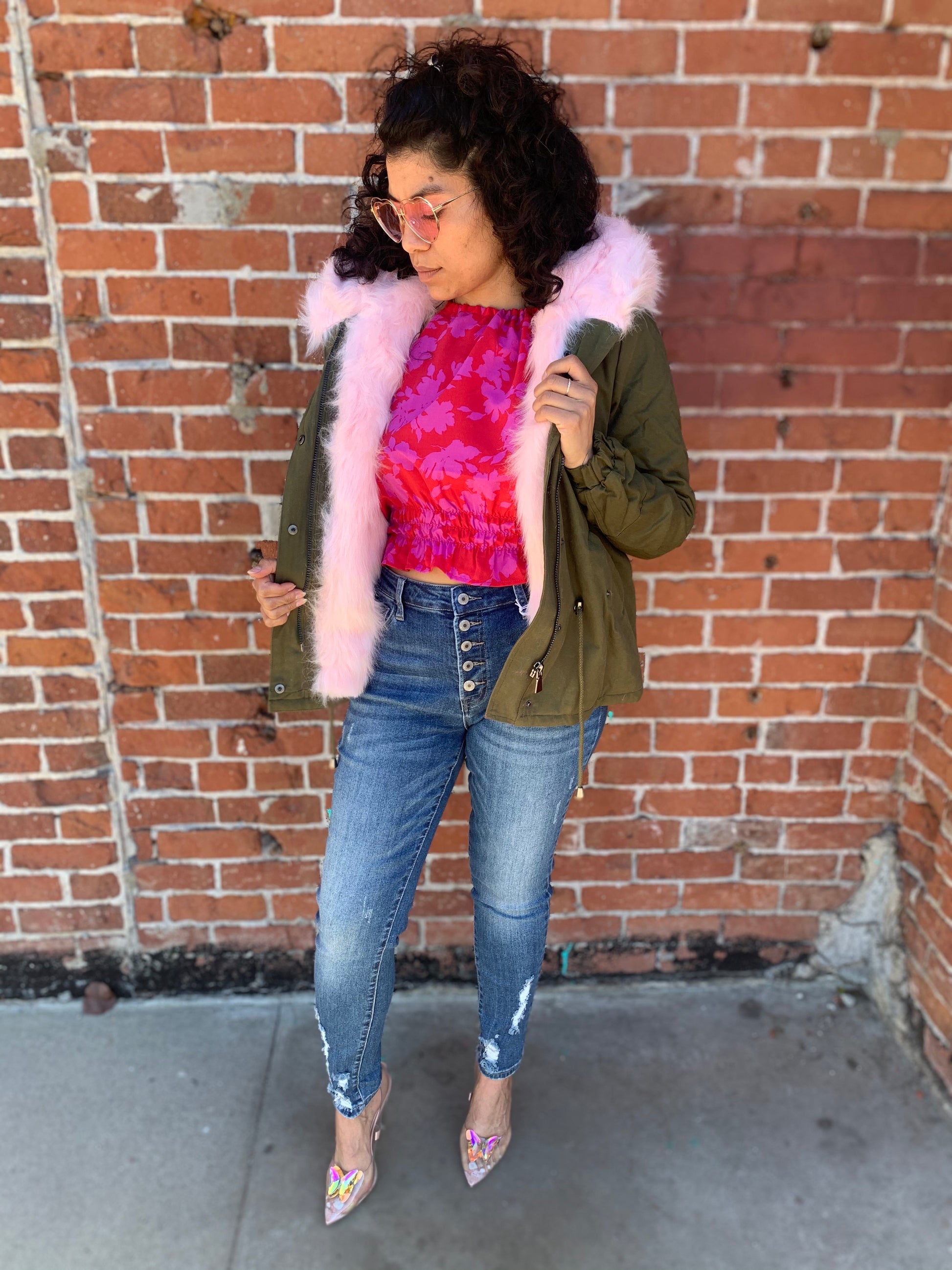model wearing an olive cargo jacket lined with pink faux fur. paired with skinny Demin jeans, a magenta floral blouse and butterfly high heels.